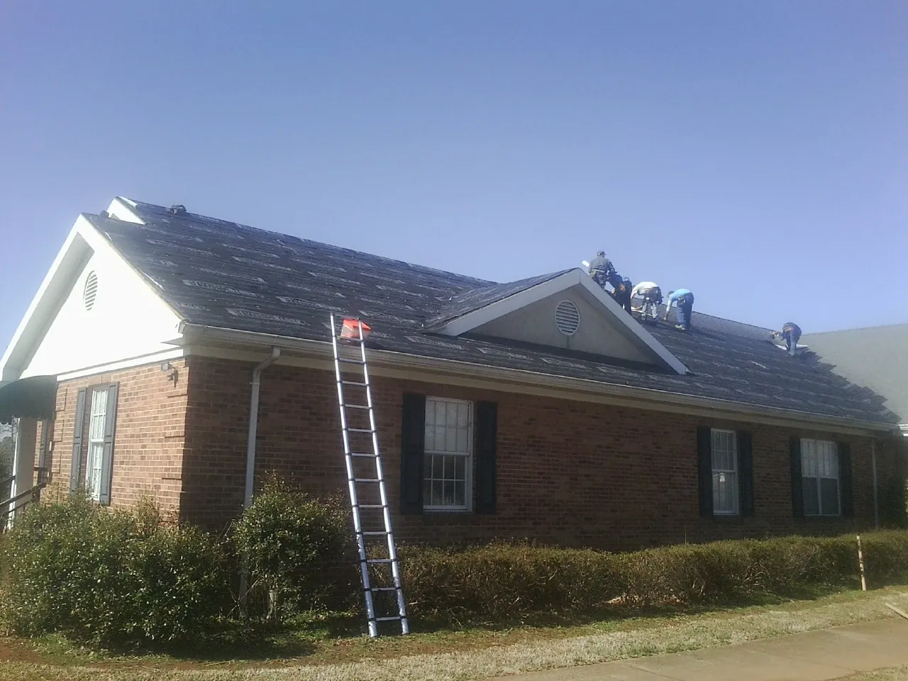 Crew tearing shingles off a brick ranch roof with a ladder set — Emergency Roof Repair work by our crew in Bellefontaine